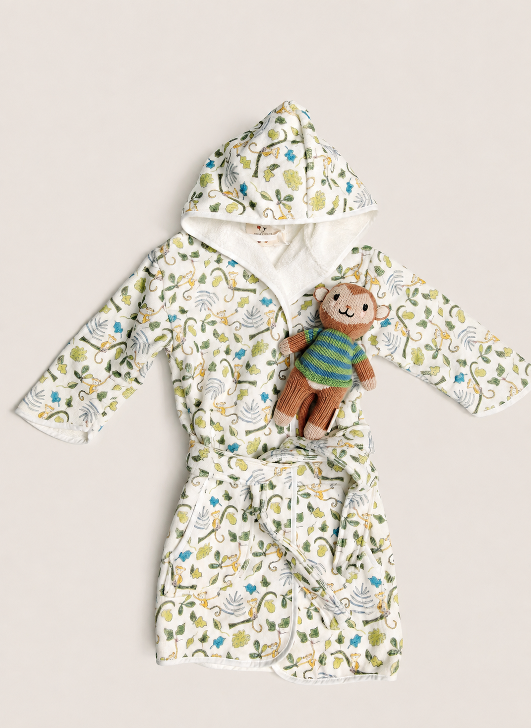 A baby hooded bathrobe with a colorful dinosaur print, styled with a small plush monkey toy, laid flat on a light beige background.