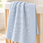 A light blue knitted baby blanket with a geometric openwork pattern draped over the side of a light wood crib in a softly lit nursery.