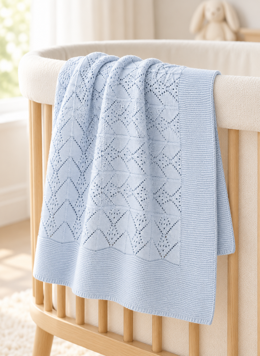 A light blue knitted baby blanket with a geometric openwork pattern draped over the side of a light wood crib in a softly lit nursery.