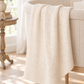 An off-white cream cable-knit baby blanket with a geometric eyelet pattern draped over the side of a light-colored wooden crib in a bright, sunlit nursery.
