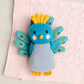 A knitted bird toy with a blue body, yellow crest and feet, and teal peacock-style wings, placed on a light pink textured blanket against a light gray background.