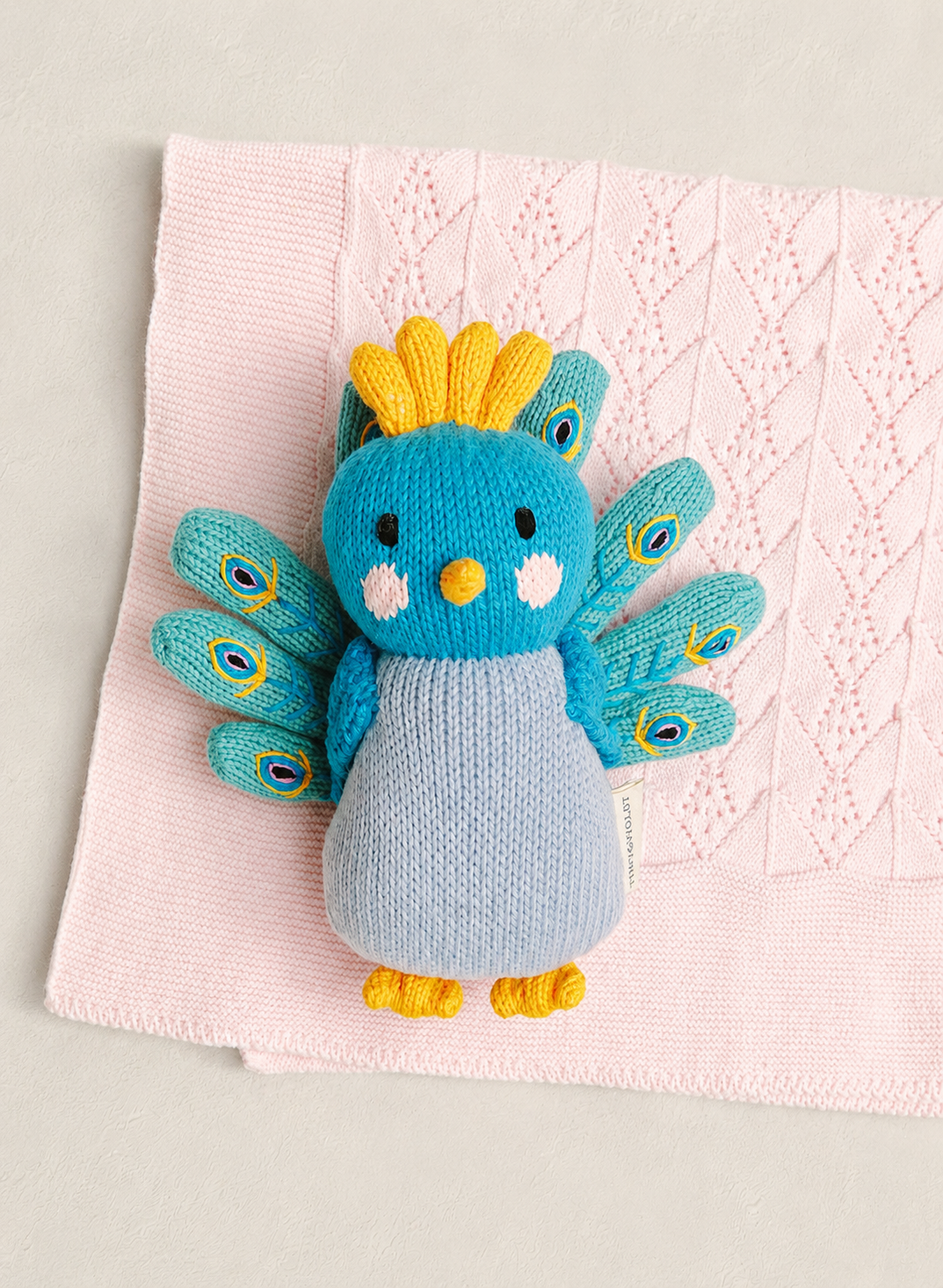 A knitted bird toy with a blue body, yellow crest and feet, and teal peacock-style wings, placed on a light pink textured blanket against a light gray background.