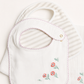 A white baby bib with pink scalloped trim and embroidered red flowers with green stems, layered over a white and beige striped muslin cloth.