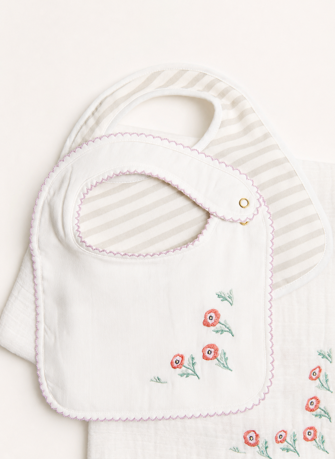 A white baby bib with pink scalloped trim and embroidered red flowers with green stems, layered over a white and beige striped muslin cloth.