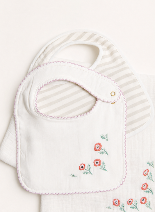 A white baby bib with pink scalloped trim and embroidered red flowers with green stems, layered over a white and beige striped muslin cloth.