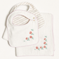 A baby gift set featuring a white cotton bib with pink trim and embroidered red flowers, layered over a matching white muslin cloth with a striped inner bib insert.
