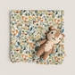 A square fabric item with an all-over woodland animal print in muted orange, green, blue, and brown tones, styled flat on a light beige background with a small plush stuffed animal placed on top.