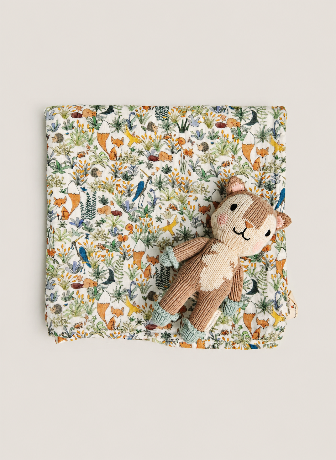 A square fabric item with an all-over woodland animal print in muted orange, green, blue, and brown tones, styled flat on a light beige background with a small plush stuffed animal placed on top.