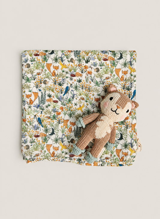 A square fabric item with an all-over woodland animal print in muted orange, green, blue, and brown tones, styled flat on a light beige background with a small plush stuffed animal placed on top.