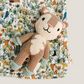 A knitted plush deer toy with a woodland-themed fabric blanket underneath, featuring a detailed pattern of trees, birds, and foxes on a light background.