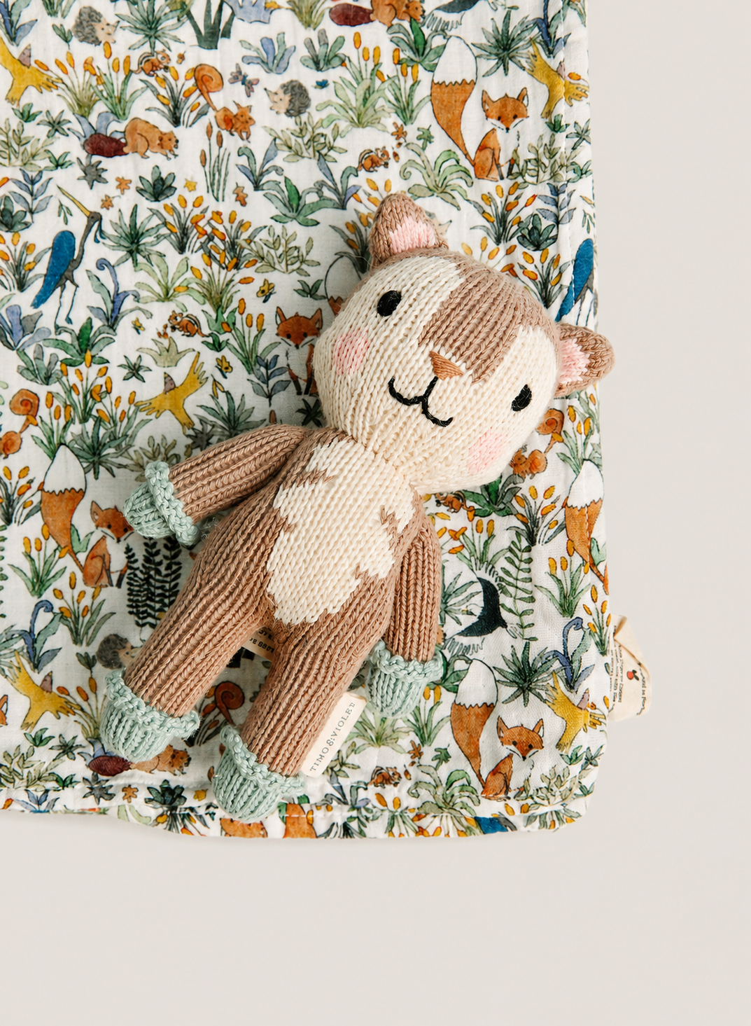 A knitted plush deer toy with a woodland-themed fabric blanket underneath, featuring a detailed pattern of trees, birds, and foxes on a light background.