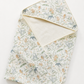 A folded baby blanket or swaddle with a delicate woodland print featuring deer, rabbits, and mushrooms on a cream background, with a solid cream inner lining and trim.