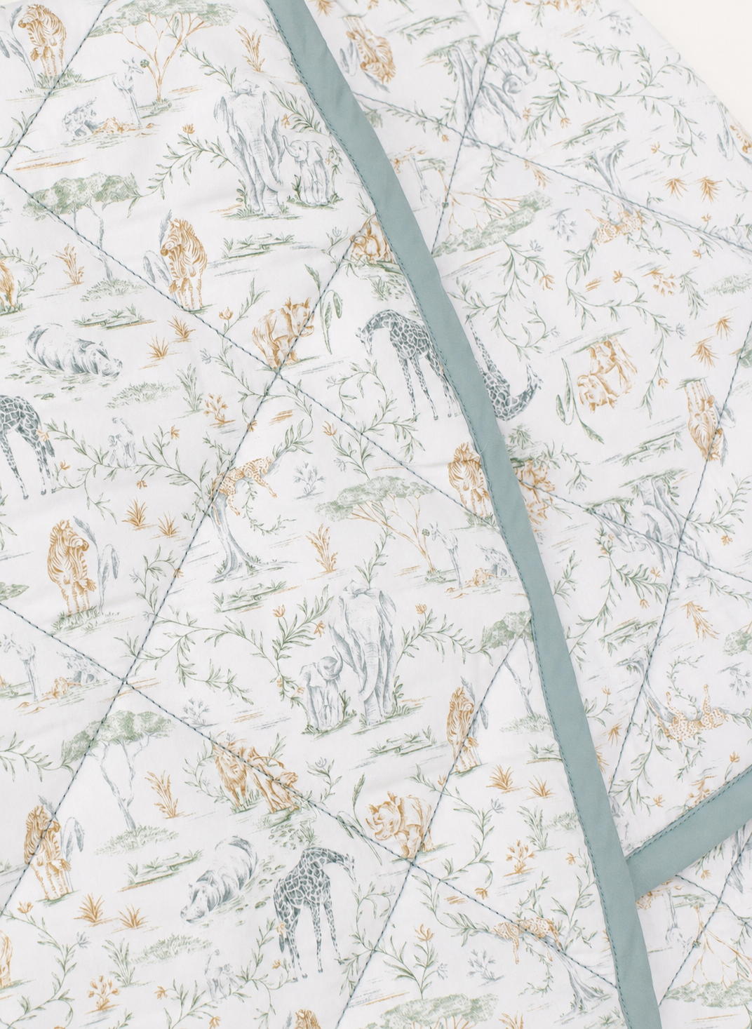 A close-up of a quilted baby blanket featuring a safari animal print with giraffes, lions, and zebras in muted blue, green, and beige tones on a white background, accented by a dusty blue-green border.