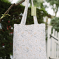 A reusable fabric tote bag with a delicate Safari pattern, hanging from a tree branch outdoors. The bag features a white base with light blue and beige floral motifs and has a light green hangtag attached.