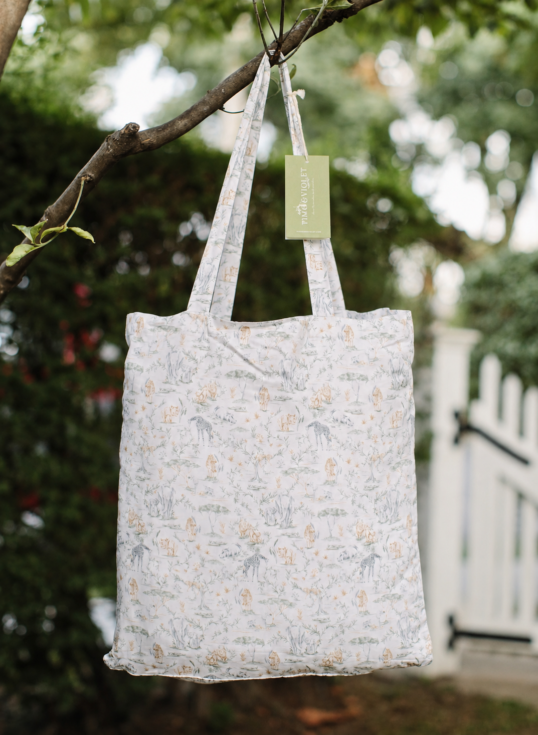 A reusable fabric tote bag with a delicate Safari pattern, hanging from a tree branch outdoors. The bag features a white base with light blue and beige floral motifs and has a light green hangtag attached.