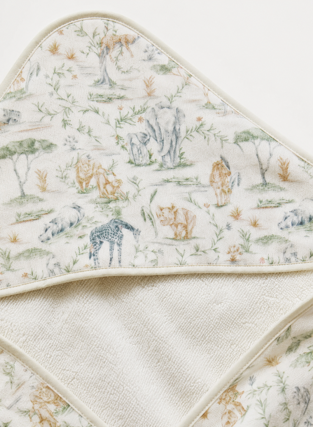 A close-up of a hooded baby towel or wrap with a soft safari animal print on an off-white background. The print features illustrated giraffes, lions, zebras, and trees in muted beige, brown, and gray tones.