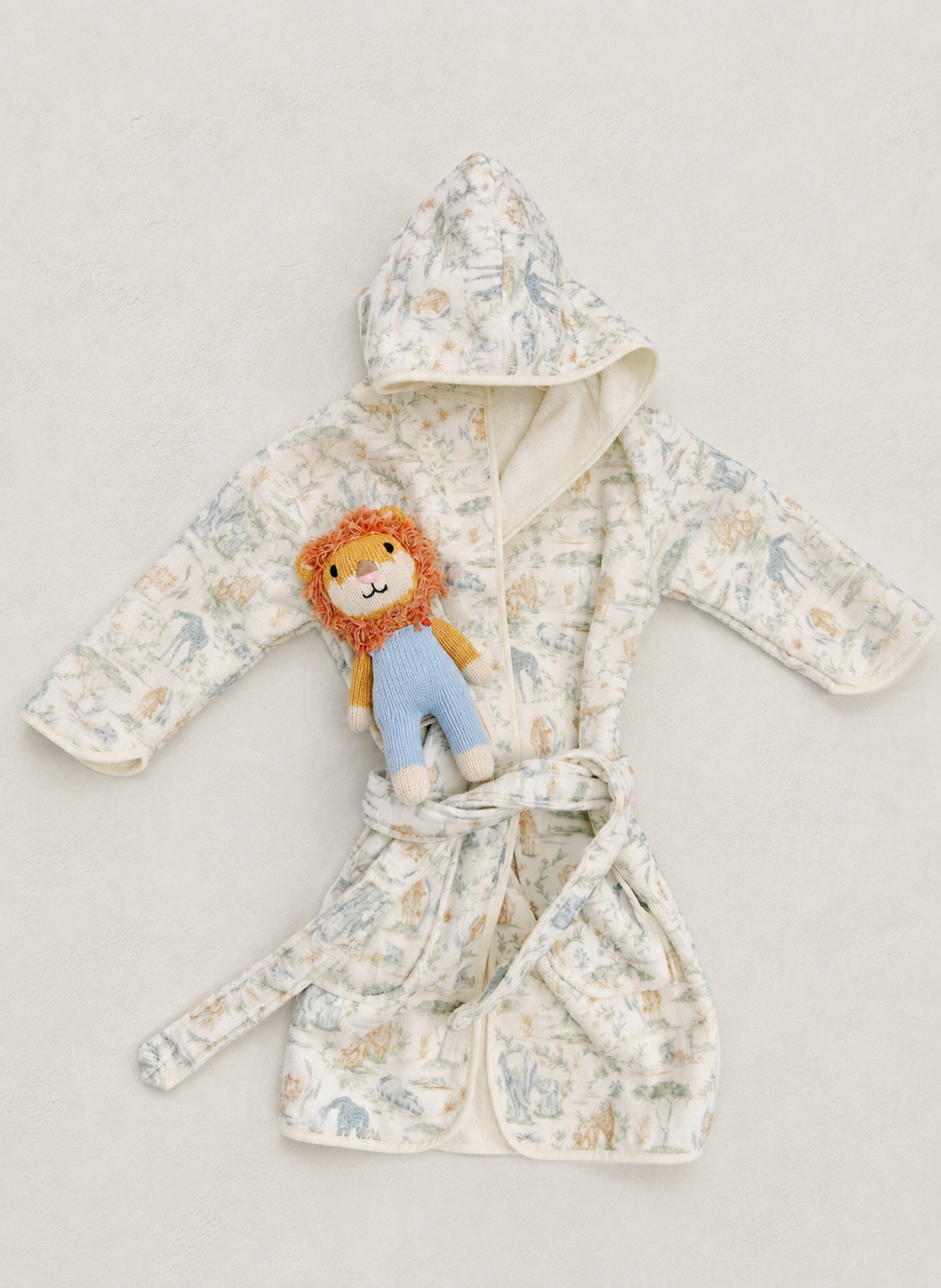 A baby hooded bathrobe with a soft cream background and a light blue and beige animal print, styled with a plush lion toy on the front, set against a neutral light beige background.