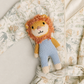 A plush lion toy with a fluffy orange-brown mane, cream face, and light blue knitted pants, laid on top of a cream-colored baby robe with a delicate floral print.
