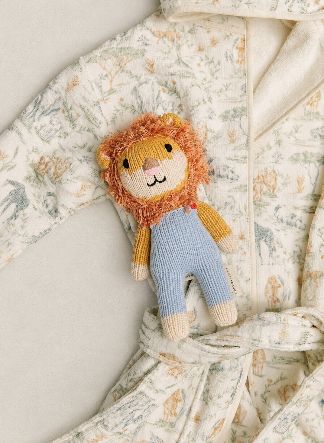 A plush lion toy with a fluffy orange-brown mane, cream face, and light blue knitted pants, laid on top of a cream-colored baby robe with a delicate floral print.