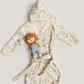 A baby hooded bathrobe with a tie belt, featuring a delicate multicolor animal print on a cream background. A small plush lion toy is tucked into the front pocket of the robe.