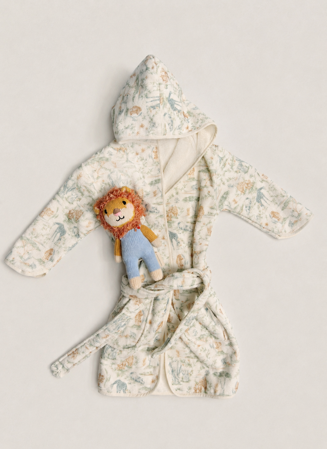 A baby hooded bathrobe with a tie belt, featuring a delicate multicolor animal print on a cream background. A small plush lion toy is tucked into the front pocket of the robe.