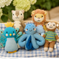 A group of small knitted animal toys arranged on a blue-and-white gingham cloth, with a soft blurred background of green leaves and pale flowers. The toys include a turquoise bird with yellow accents, a light blue octopus, a brown monkey in a green-and-blue striped shirt, a tan bear, a lion with a tan mane, and a gray cat.