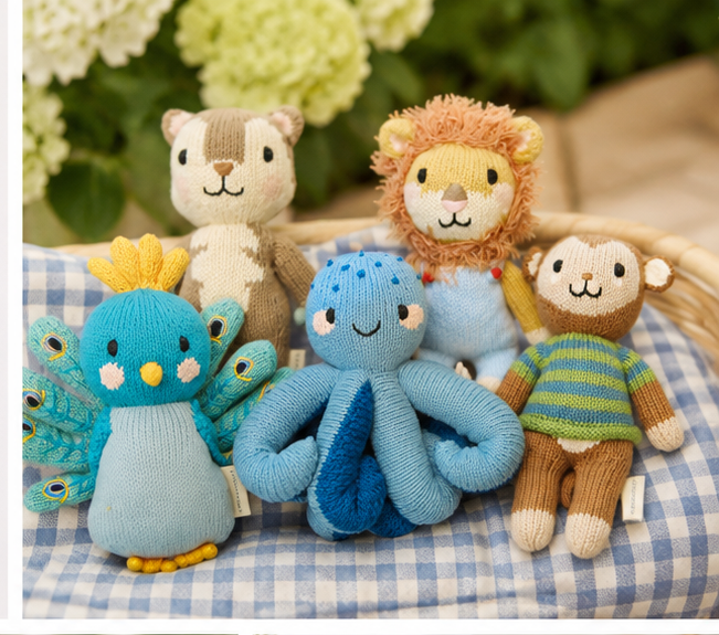 A group of small knitted animal toys arranged on a blue-and-white gingham cloth, with a soft blurred background of green leaves and pale flowers. The toys include a turquoise bird with yellow accents, a light blue octopus, a brown monkey in a green-and-blue striped shirt, a tan bear, a lion with a tan mane, and a gray cat.
