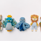 Five knitted plush baby toys arranged in a row on a white background: a brown and cream bear, a blue peacock with yellow accents, a blue octopus, a lion in yellow and blue, and a brown monkey in a green striped shirt.