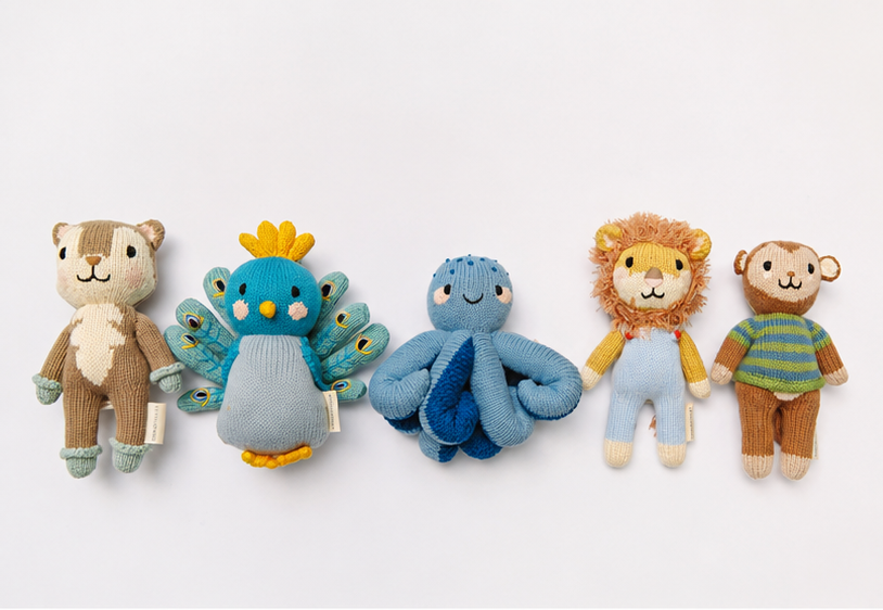 Five knitted plush baby toys arranged in a row on a white background: a brown and cream bear, a blue peacock with yellow accents, a blue octopus, a lion in yellow and blue, and a brown monkey in a green striped shirt.