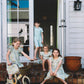 Four children wearing matching floral print dresses and shorts sit and ride on a porch in front of a white house. One girl rides a cream bicycle with a basket, while the others sit on the steps and in the doorway.