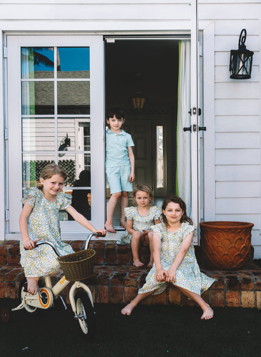 Four children wearing matching floral print dresses and shorts sit and ride on a porch in front of a white house. One girl rides a cream bicycle with a basket, while the others sit on the steps and in the doorway.