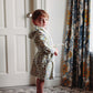 A young child wearing a cream hooded robe with a blue, green, and yellow floral pattern, standing barefoot in a room with a white door, patterned curtains, and a geometric rug.