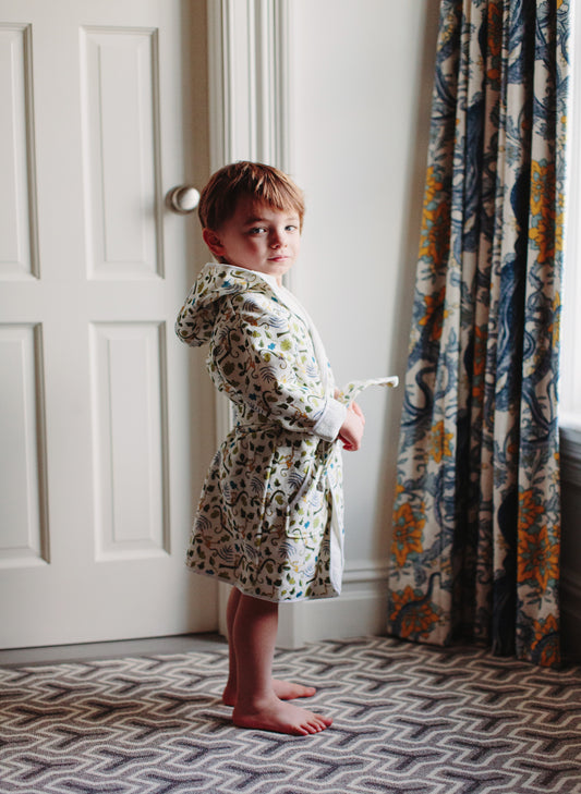 A young child wearing a cream hooded robe with a blue, green, and yellow floral pattern, standing barefoot in a room with a white door, patterned curtains, and a geometric rug.