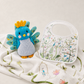 A styled flat-lay featuring a blue and gray knitted bird plush toy with yellow accents, next to a white baby bib with colorful embroidered birds and floral patterns, all resting on a matching white embroidered blanket.