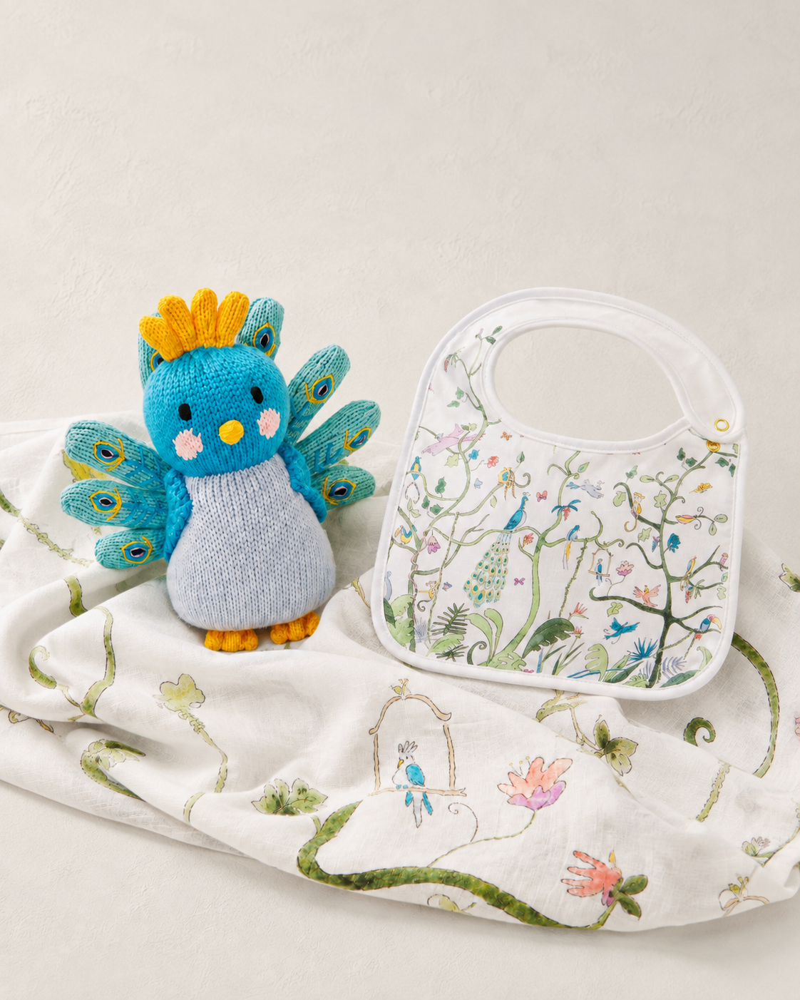 A styled flat-lay featuring a blue and gray knitted bird plush toy with yellow accents, next to a white baby bib with colorful embroidered birds and floral patterns, all resting on a matching white embroidered blanket.