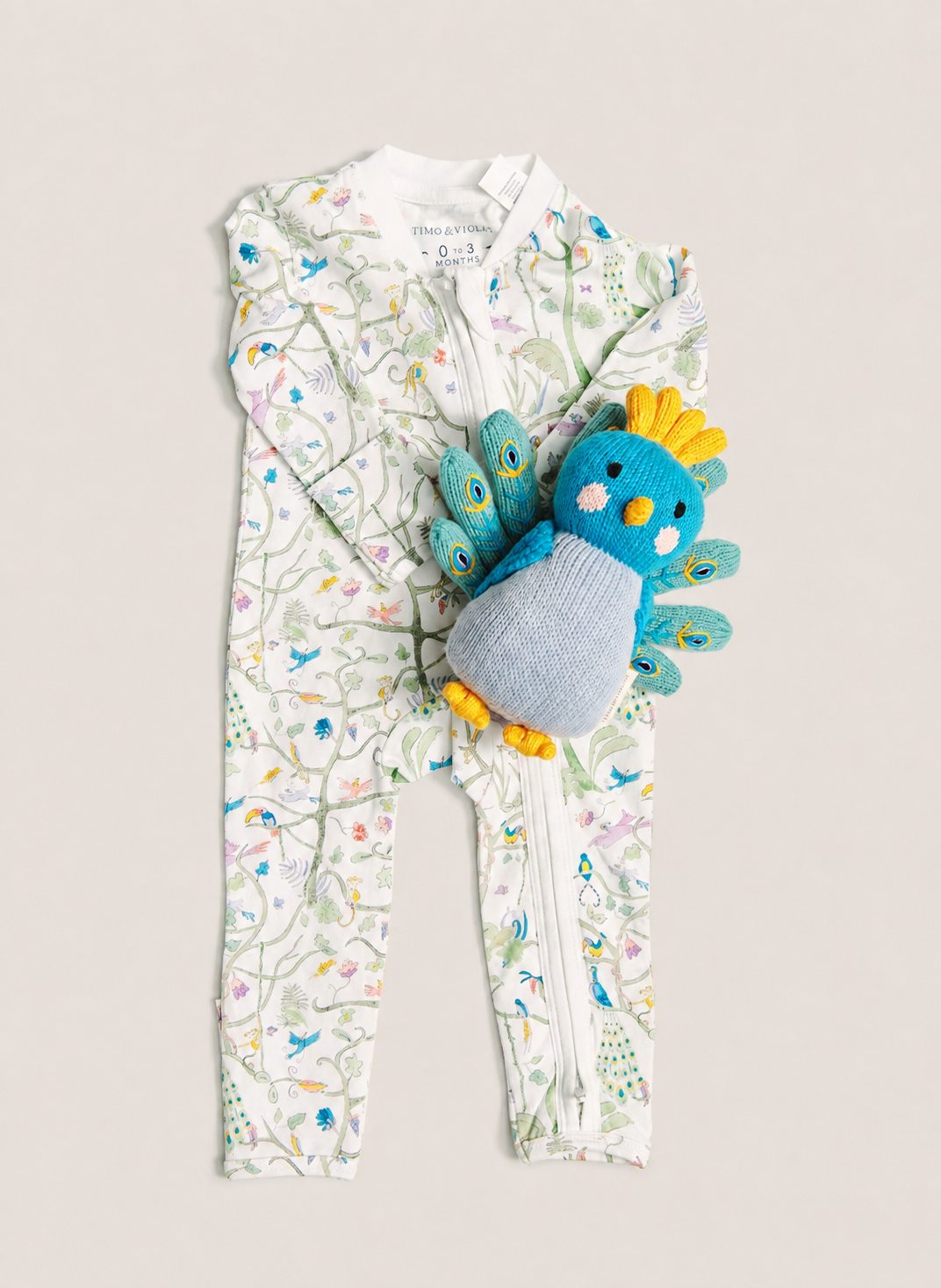A baby's full-zip footless one-piece romper in white with a colorful bird and floral print, styled with a blue and yellow plush toy chick on a light beige background.