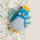 A knitted bird-shaped baby accessory with a blue head, yellow crown and feet, and light blue body, placed on top of a floral printed baby garment.