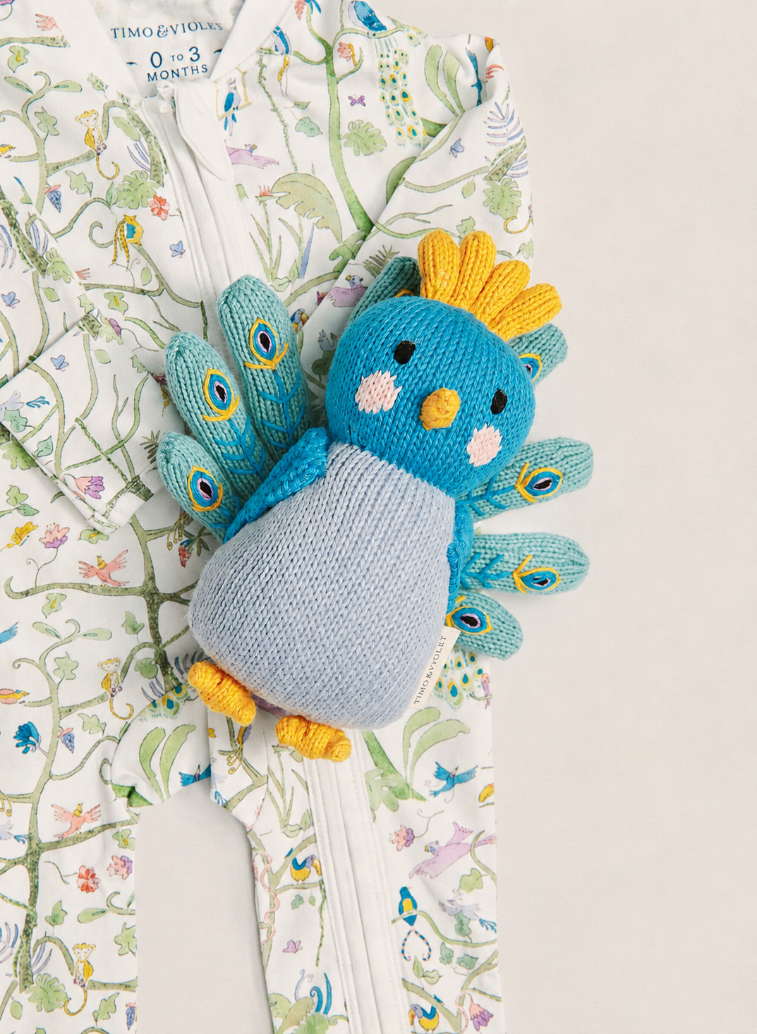 A knitted bird-shaped baby accessory with a blue head, yellow crown and feet, and light blue body, placed on top of a floral printed baby garment.