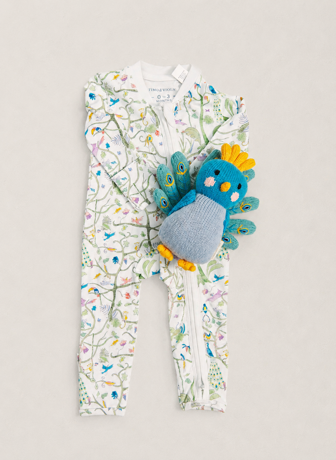 A baby's footed zip-up sleeper with a white background and a colorful bird and floral print, styled with a blue and yellow plush bird toy on a light beige background.