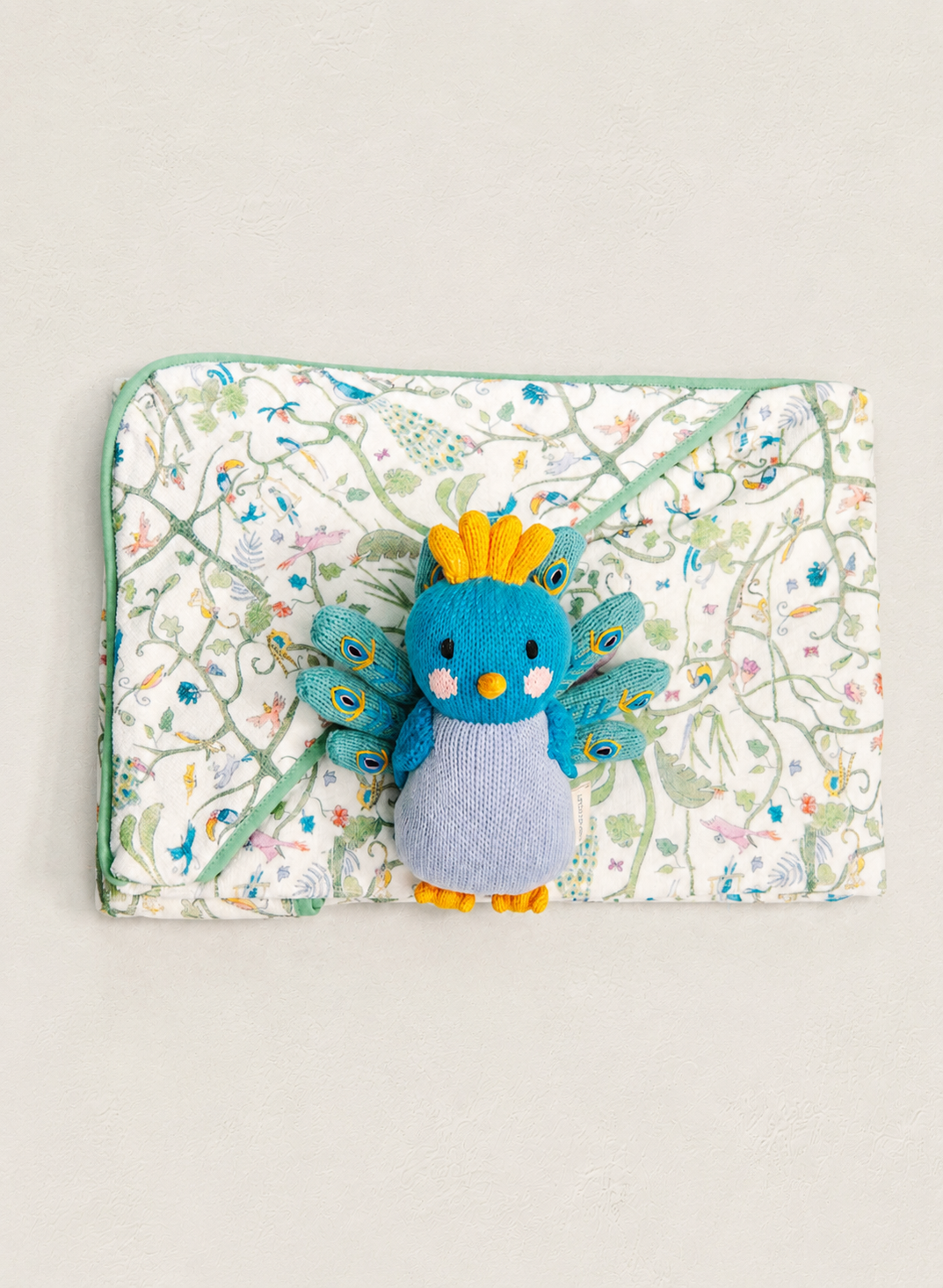 A blue and gray plush bird toy with yellow accents is placed on top of a square quilted fabric pouch featuring a delicate multicolor floral and vine pattern on a white background.