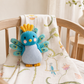 A small knitted bird toy in blue, teal, yellow, and light blue sits on a cream muslin baby blanket draped over a wooden crib in a softly lit nursery.