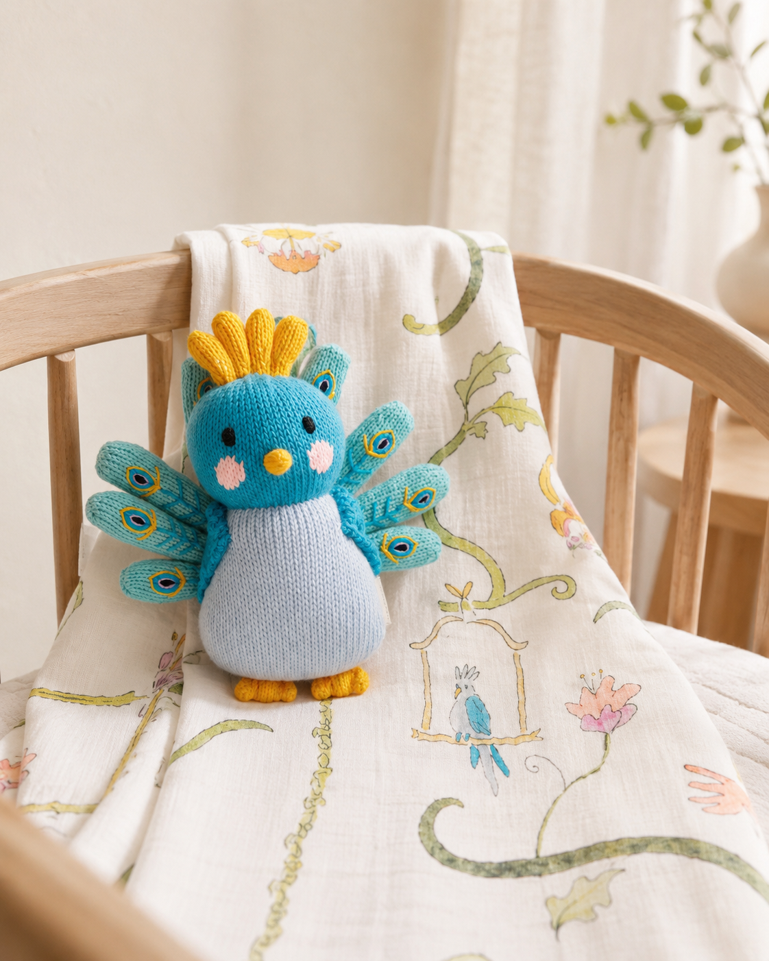 A small knitted bird toy in blue, teal, yellow, and light blue sits on a cream muslin baby blanket draped over a wooden crib in a softly lit nursery.
