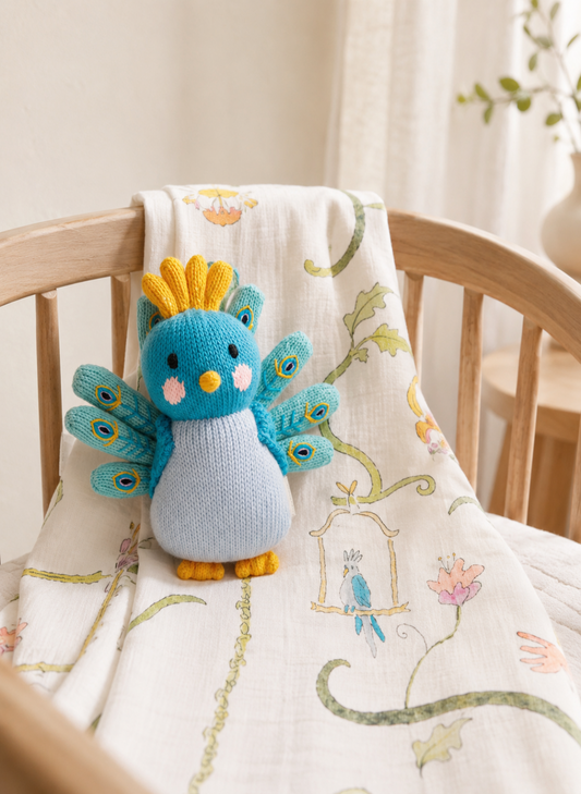 A small knitted bird toy in blue, teal, yellow, and light blue sits on a cream muslin baby blanket draped over a wooden crib in a softly lit nursery.