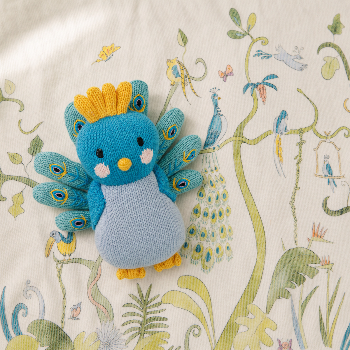 A knitted peacock bird toy in shades of blue, yellow, and light gray, placed on a cream-colored fabric background with a botanical and bird pattern.