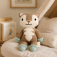 A knitted plush toy animal with a brown and cream body, black embroidered facial features, and mint green accents on its paws and cuffs, sitting on a beige cushion in a cozy room setting with books and framed art in the background.
