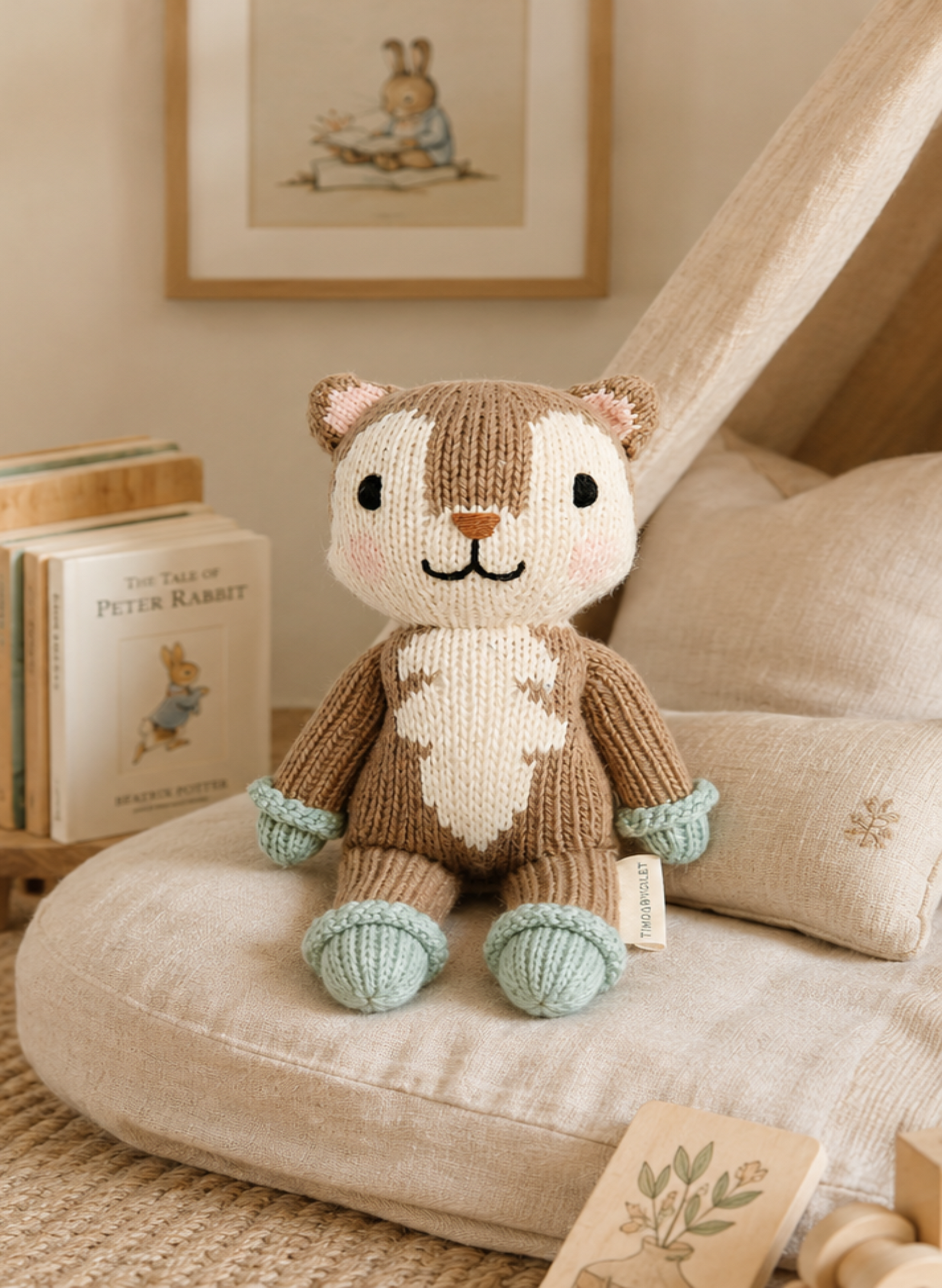 A knitted plush toy animal with a brown and cream body, black embroidered facial features, and mint green accents on its paws and cuffs, sitting on a beige cushion in a cozy room setting with books and framed art in the background.
