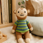 A knitted plush monkey toy wearing a striped sweater in brown, green, and blue, posed on a soft cream fabric with a bookshelf, basket, and plant in the background.