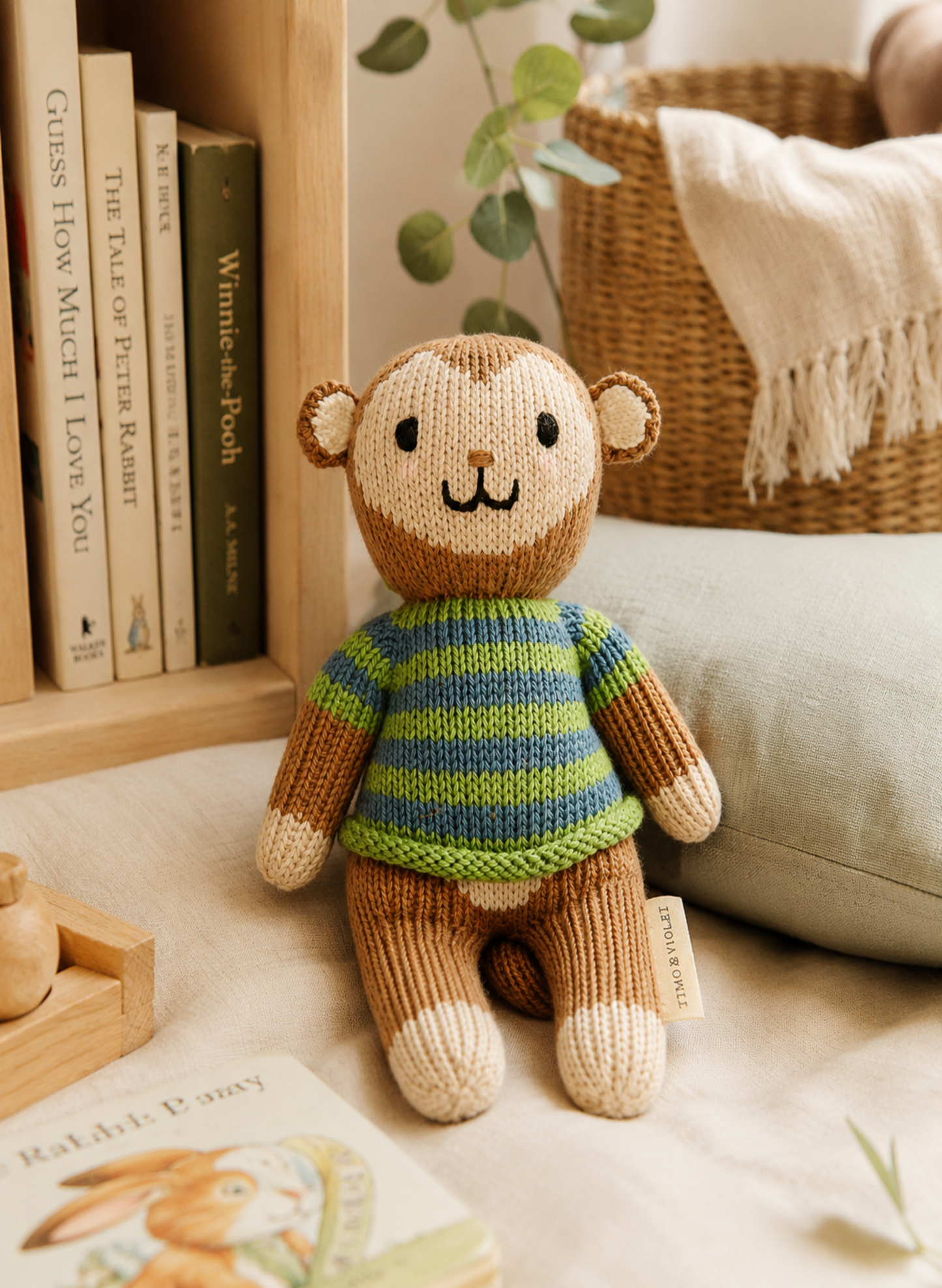 A knitted plush monkey toy wearing a striped sweater in brown, green, and blue, posed on a soft cream fabric with a bookshelf, basket, and plant in the background.