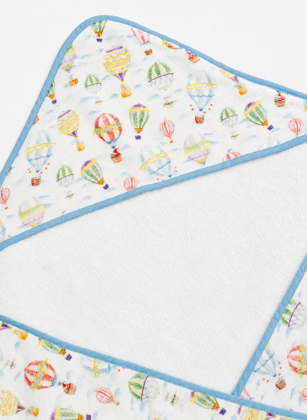 A close-up of a hooded baby towel showing the printed outer fabric with colorful hot air balloons on a white background and a light blue trim, alongside the soft white plush inner lining.
