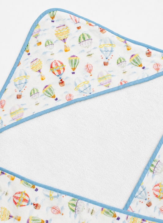 A close-up of a hooded baby towel showing the printed outer fabric with colorful hot air balloons on a white background and a light blue trim, alongside the soft white plush inner lining.